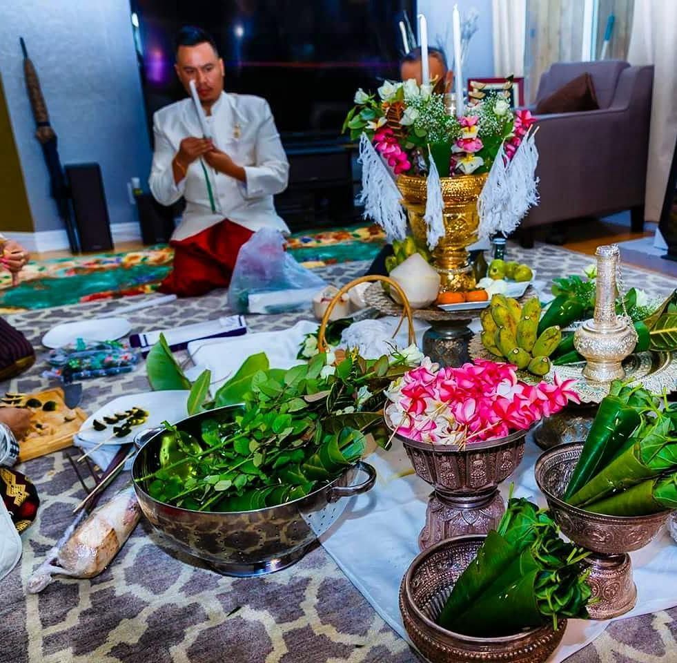 A baci setup on the floor of a home — the pha khuan hung with flowers and white threads, bowls of leaves and plumeria, silver vessels, Thip in white preparing the offerings behind it.