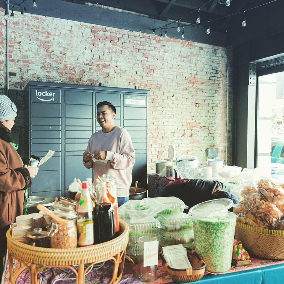 Thip laughing behind a market table crowded with jars, sauces, bags of crackling and stacked containers, a customer in front of him.