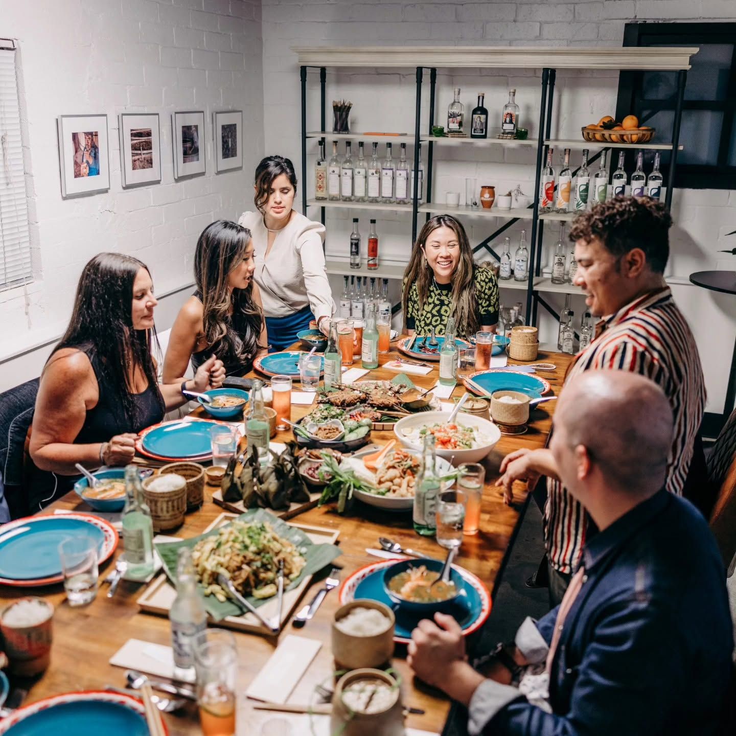 A long wooden table crowded with Lao dishes — noodle soups, grilled skewers, salads, sticky rice baskets, banana-leaf parcels — and eight people leaning in, talking, eating.